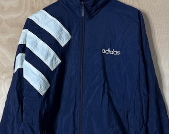 Könnte beinhalten: Marineblaue Adidas-Trainingsjacke mit weißen Streifen auf der linken Schulter und dem Ärmel. Die Jacke hat einen durchgehenden Reißverschluss, zwei Taschen und das Adidas-Logo auf der Brust. Die Jacke hängt an einem Holzbügel.
