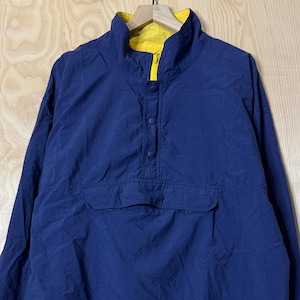 May include: A navy blue anorak jacket with a yellow lining. The jacket features a high collar, buttoned placket, and a front pocket. It is suspended from a black hook.
