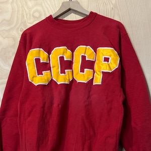 Peut inclure: Un sweat-shirt rouge vintage avec les lettres "CCCP" jaunes, bordées de blanc. Le sweat-shirt à manches longues a des poignets et un ourlet côtelés. Le vêtement est suspendu à un cintre en bois.