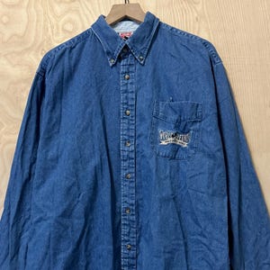 May include: A long-sleeved, button-down denim shirt in a medium blue wash. The shirt has a button-down collar, a chest pocket with embroidered text, and buttoned cuffs. The shirt is hanging on a wooden hanger.