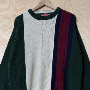 May include: A long-sleeve crewneck sweater featuring a color-blocked design in dark green, cream, and a vertical panel of maroon and navy blue. The cream section includes a vertical cable knit pattern, while the rest of the sweater is smooth knit.