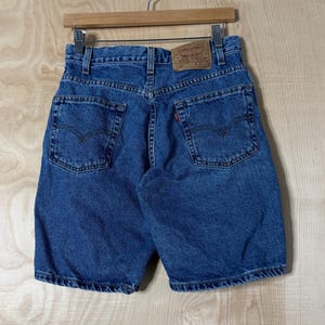 May include: Blue denim shorts featuring a high-waisted design with a tan leather patch on the back waistband, two back pockets with stitched detailing, a small red tab on the right pocket, and a button fly closure, all hanging on a wooden hanger.