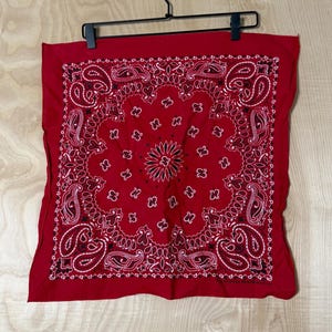 Może przedstawiać: Kwadratowa czerwona bandana ze szczegółowym czarno-białym wzorem paisley. Projekt zawiera centralny motyw przypominający kwiat, otoczony skomplikowanymi zawijasami i geometrycznymi obramowaniami, nadrukowany na jednolitym czerwonym materiale.