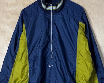 May include: A navy blue and yellow Nike windbreaker jacket with a half-zip closure. The jacket features a small white Nike swoosh logo on the chest and a striped collar. The jacket is hanging on a black hook.