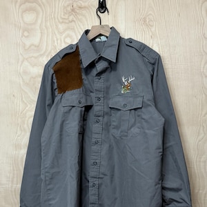 May include: A gray long-sleeved button-up shirt with a brown suede patch on the left shoulder. The shirt has a deer embroidered on the right chest.