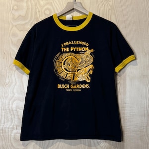 Pode incluir: Camiseta preta com detalhes amarelos. Apresenta a frase "I Challenged The Python" acima de um gráfico de cobra. Inclui também o texto "Dusch Gardens, Tampa, Florida".