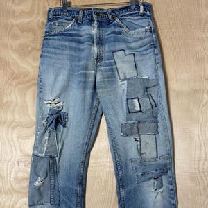 May include: Light blue denim jeans with multiple patches of varying shades of blue. The jeans have distressed areas and are hanging on a wooden hanger. The patches are rectangular and square.