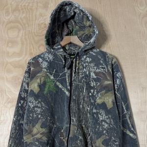 May include: A long-sleeve zip-up hoodie featuring a realistic forest camouflage pattern in shades of dark grey, black, and muted green. The garment includes a hood with black drawstrings, a full-length front zip, and two front hand pockets.