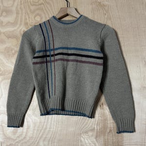 May include: A tan sweater with a crew neck and long sleeves. The sweater features a striped design across the chest in blue, black, and burgundy. The cuffs and hem are trimmed with a thin blue band. The sweater is hanging on a wooden hanger.