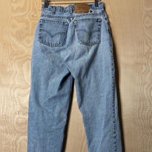 May include: Light wash denim jeans hanging on a wooden hanger. The jeans have two back pockets with a curved stitch design and a brown leather patch. The jeans are a classic style with a straight leg.