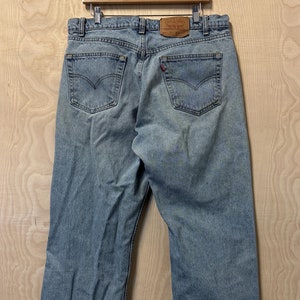 Könnte beinhalten: Hellblaue Jeans aus Denim mit ausgefransten Säumen, die an einem Holzbügel vor einem hellbraunen Hintergrund hängen. Die Jeans haben ein braunes Leder-Etikett mit dem Schriftzug "Levi's" auf der Rückseite.