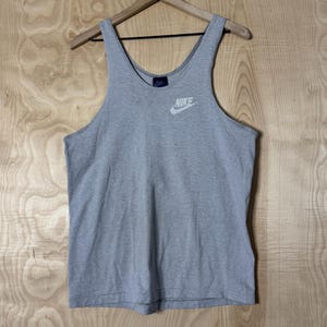 Puede incluir: Camiseta sin mangas Nike gris con el logo blanco de Nike en el pecho. La camiseta sin mangas está colgada en una percha de madera sobre un fondo de madera clara. La camiseta sin mangas tiene un estilo atlético clásico.