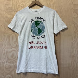Peut inclure: T-shirt blanc avec un motif de globe terrestre et l'inscription "WE CHANGE THE WORLD". En dessous, on peut lire "GIRL SCOUTS LAKAMAGA '91" en rouge. Le t-shirt est suspendu &agrave; un cintre en bois.