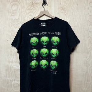 Puede incluir: Camiseta negra con un gráfico de un extraterrestre verde. El gráfico muestra nueve caras de extraterrestres diferentes con diferentes expresiones, cada una etiquetada con un estado de ánimo: Feliz, Triste, Enojado, Cansado, Confundido, Asustado, Hambriento, Perdido y Acosado.