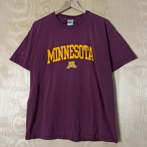 May include: A maroon short-sleeved t-shirt featuring the word MINNESOTA in bold yellow lettering across the chest, positioned above a yellow block letter M logo. The shirt has a crew neckline and is displayed on a wooden hanger against a wall.