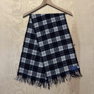 Pode incluir: Uma echarpe xadrez preta e branca com uma borda franjada. A echarpe está pendurada em um cabide de madeira.