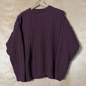 May include: A deep purple cable-knit sweater hangs on a wooden hanger. The sweater features a crew neck and a thick, textured knit pattern. The sleeves are wide and the overall style is oversized.