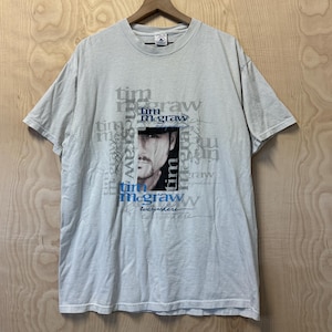 Puede incluir: Una camiseta gris claro con un retrato de Tim McGraw. La camiseta tiene el texto "Tim McGraw" en un patrón repetido y la palabra "everywhere" debajo del retrato. La camiseta está colgada en una percha de madera.