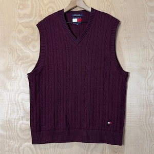 May include: A burgundy cable-knit sweater vest with a V-neck. The vest has a small logo patch on the lower right and a brand tag at the back of the neck. The vest is hanging on a wooden hanger against a light wood background.