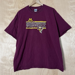 Peut inclure: T-shirt bordeaux avec l'inscription "UNIVERSITY OF MINNESOTA FOOTBALL" en lettres dorées et blanches. Le t-shirt a des manches courtes et un col rond. Un petit graphisme d'un joueur de football est également visible.
