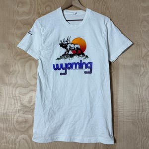 May include: White t-shirt featuring a graphic of a moose silhouette against a setting sun, with the word "Wyoming" in blue and orange. The shirt is short-sleeved and hangs on a wooden hanger.