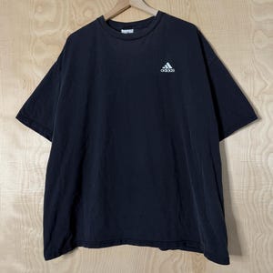 May include: Black t-shirt with a small white Adidas logo on the upper left chest. The shirt is hanging on a wooden hanger against a light wood grain background. The t-shirt has short sleeves and a crew neck.
