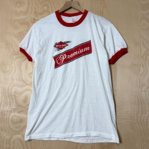 May include: A white ringer t-shirt with red trim on the collar and sleeve cuffs. The front features a red diagonal banner with the word Premium in white script, and a small red diamond logo above it with the text Grain Belt Beer.