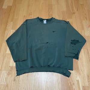 May include: A green sweatshirt with a distressed look, featuring a small Nike logo on the left chest. The sweatshirt has several holes and frayed edges.