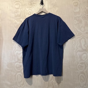 May include: A navy blue short-sleeved t-shirt hanging on a wooden hanger. The shirt is made of a soft material and has a relaxed fit. The background is a light-coloured wooden wall.