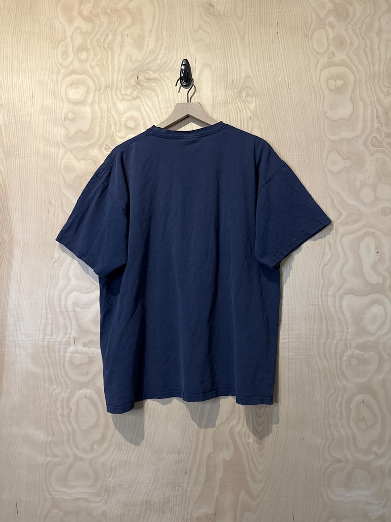 May include: A navy blue short-sleeved t-shirt hanging on a wooden hanger. The shirt is made of a soft material and has a relaxed fit. The background is a light-coloured wooden wall.