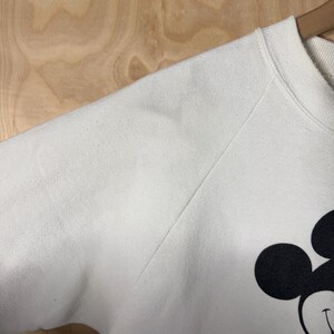 May include: White sweatshirt featuring a large graphic of Mickey Mouse. The sweatshirt has a crew neck and long sleeves. The Mickey Mouse graphic includes black, red, yellow, and white colors. The sweatshirt is hanging on a wooden hanger.