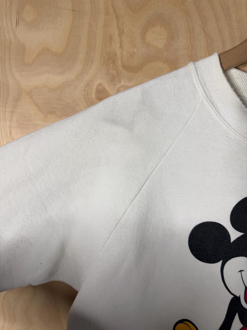 May include: White sweatshirt featuring a large graphic of Mickey Mouse. The sweatshirt has a crew neck and long sleeves. The Mickey Mouse graphic includes black, red, yellow, and white colors. The sweatshirt is hanging on a wooden hanger.