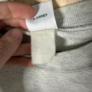 K&ouml;nnte beinhalten: Nahaufnahme eines grauen Sweatshirts mit einem Etikett mit der Aufschrift "&copy; DISNEY". Das Etikett ist wei&szlig; mit schwarzem Text. Das Sweatshirt besteht aus einem weichen, strukturierten Stoff.