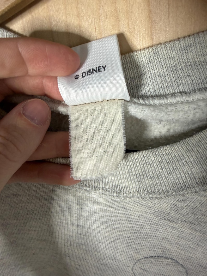 K&ouml;nnte beinhalten: Nahaufnahme eines grauen Sweatshirts mit einem Etikett mit der Aufschrift "&copy; DISNEY". Das Etikett ist wei&szlig; mit schwarzem Text. Das Sweatshirt besteht aus einem weichen, strukturierten Stoff.