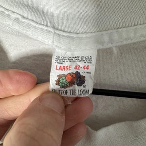 May include: Close-up of a white t-shirt tag. The tag reads "ALL COTTON MADE IN USA" and "LARGE 42-44". The tag also features the Fruit of the Loom logo with a graphic of fruit. The shirt is made of a soft, textured fabric.