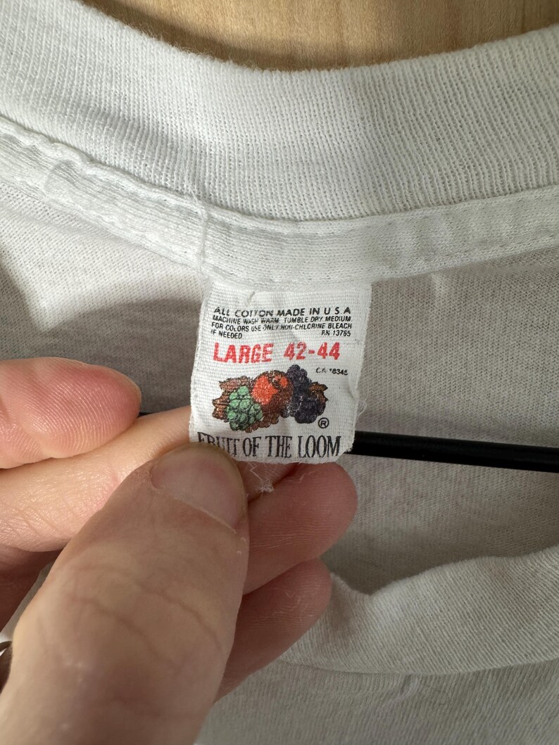 May include: Close-up of a white t-shirt tag. The tag reads "ALL COTTON MADE IN USA" and "LARGE 42-44". The tag also features the Fruit of the Loom logo with a graphic of fruit. The shirt is made of a soft, textured fabric.