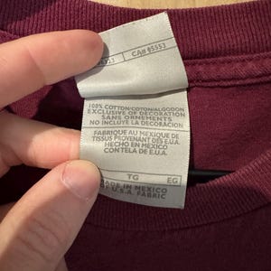 K&ouml;nnte beinhalten: Nahaufnahme eines weinroten T-Shirt-Etiketts mit Text. Das wei&szlig;e Etikett hat schwarzen Text, darunter "100% COTTON" und "MADE IN MEXICO". Das Etikett hat auch eine CA#- und Gr&ouml;&szlig;enangaben. Das T-Shirt hat eine einfarbige weinrote Farbe.