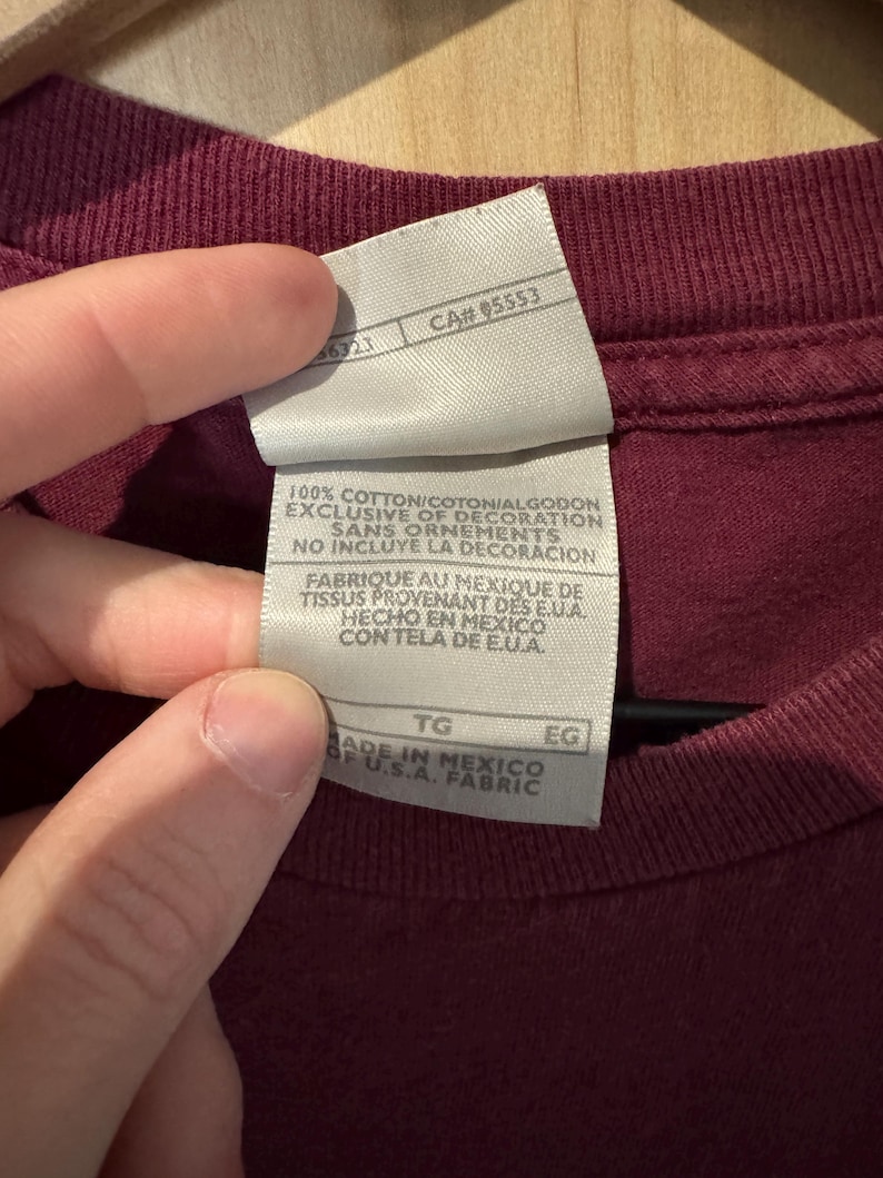 K&ouml;nnte beinhalten: Nahaufnahme eines weinroten T-Shirt-Etiketts mit Text. Das wei&szlig;e Etikett hat schwarzen Text, darunter "100% COTTON" und "MADE IN MEXICO". Das Etikett hat auch eine CA#- und Gr&ouml;&szlig;enangaben. Das T-Shirt hat eine einfarbige weinrote Farbe.