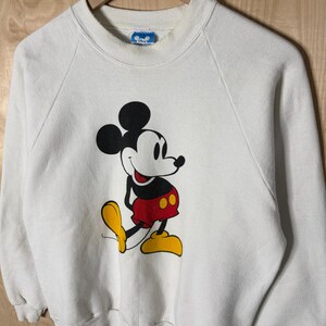May include: White sweatshirt featuring a large graphic of Mickey Mouse. The cartoon character is depicted in black, red, and yellow. The sweatshirt has a crew neck and long sleeves. The Disney logo is visible on the collar.