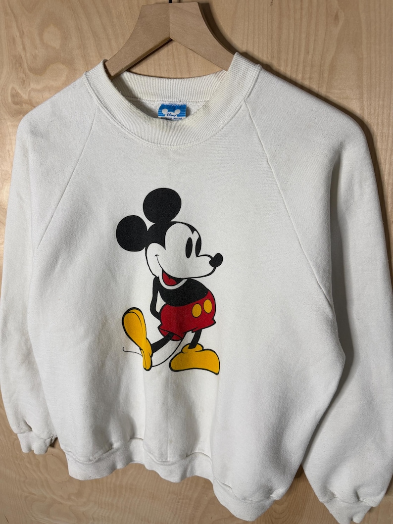May include: White sweatshirt featuring a large graphic of Mickey Mouse. The cartoon character is depicted in black, red, and yellow. The sweatshirt has a crew neck and long sleeves. The Disney logo is visible on the collar.