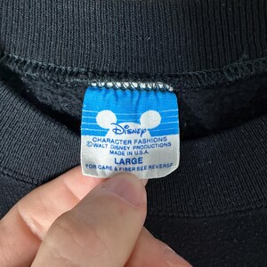 May include: Close-up of a black sweatshirt tag with the Disney logo and text. The blue tag reads "CHARACTER FASHIONS WALT DISNEY PRODUCTIONS MADE IN U.S.A. LARGE". The tag is held by a hand.