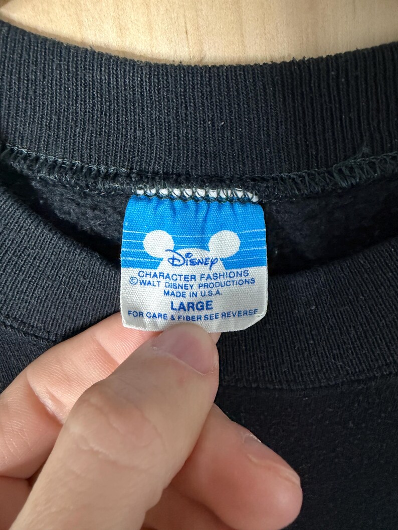 May include: Close-up of a black sweatshirt tag with the Disney logo and text. The blue tag reads "CHARACTER FASHIONS WALT DISNEY PRODUCTIONS MADE IN U.S.A. LARGE". The tag is held by a hand.