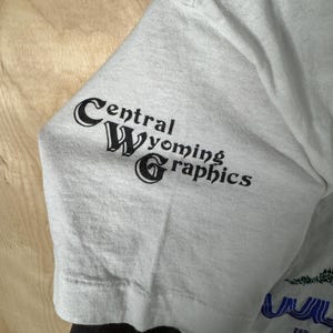 May include: A light gray t-shirt sleeve with the text "Central Wyoming Graphics" printed in black. The sleeve is made of a soft, slightly wrinkled fabric. The shirt is a light gray color.