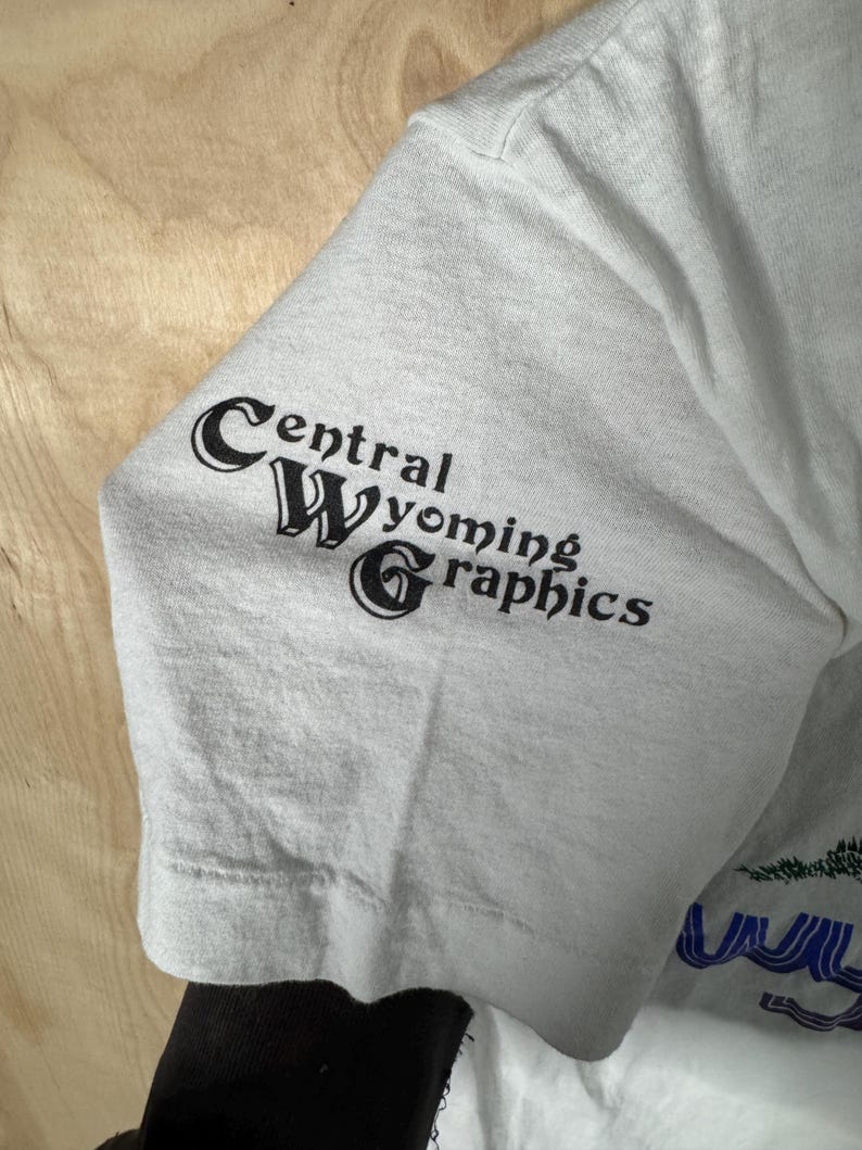 May include: A light gray t-shirt sleeve with the text "Central Wyoming Graphics" printed in black. The sleeve is made of a soft, slightly wrinkled fabric. The shirt is a light gray color.