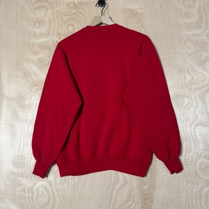 May include: A plain red sweatshirt with a crew neck and long sleeves. The cuffs and hem are ribbed. The garment is suspended on a wooden hanger against a light wood grain backdrop.