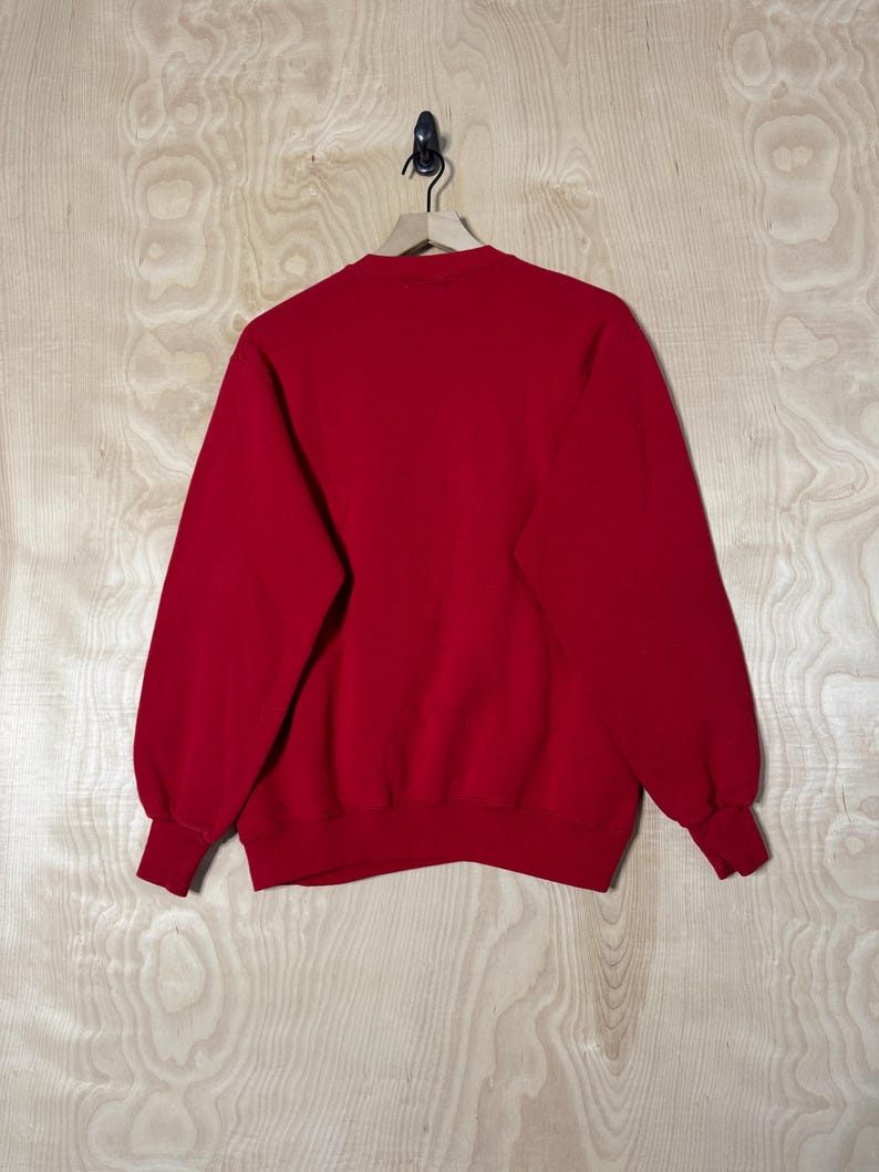May include: A plain red sweatshirt with a crew neck and long sleeves. The cuffs and hem are ribbed. The garment is suspended on a wooden hanger against a light wood grain backdrop.