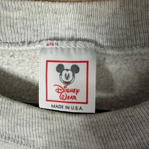K&ouml;nnte beinhalten: Nahaufnahme eines grauen Sweatshirt-Etiketts mit dem Disney Wear-Logo mit Mickey Mouse. Das Etikett ist rechteckig mit rotem Rand und dem Text "MADE IN U.S.A." ist darunter gedruckt. Das Sweatshirt hat einen gerippten Ausschnitt.