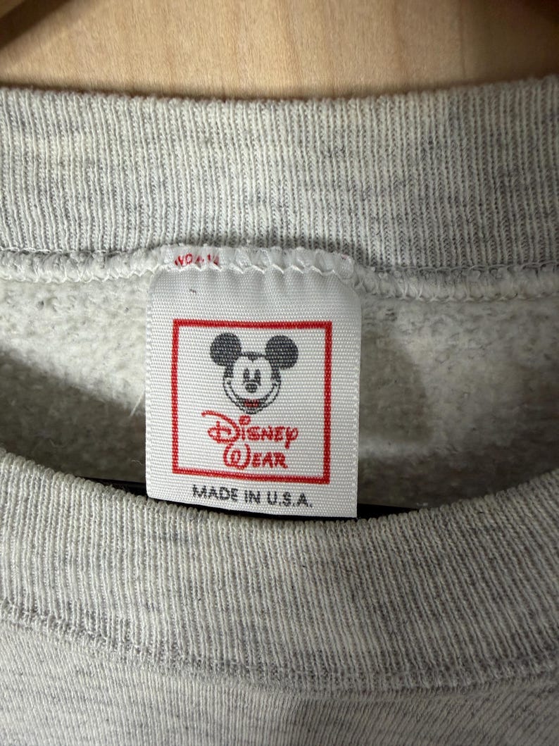 K&ouml;nnte beinhalten: Nahaufnahme eines grauen Sweatshirt-Etiketts mit dem Disney Wear-Logo mit Mickey Mouse. Das Etikett ist rechteckig mit rotem Rand und dem Text "MADE IN U.S.A." ist darunter gedruckt. Das Sweatshirt hat einen gerippten Ausschnitt.