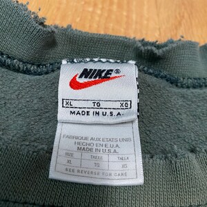 May include: A green Nike sweatshirt tag with the text "XL TO XG MADE IN U.S.A." and "FABRIQUE AUX ETATS UNIS HECHO EN E.U.A. MADE IN U.S.A.". The tag also has a size chart with the sizes "XL", "TG", and "XG".