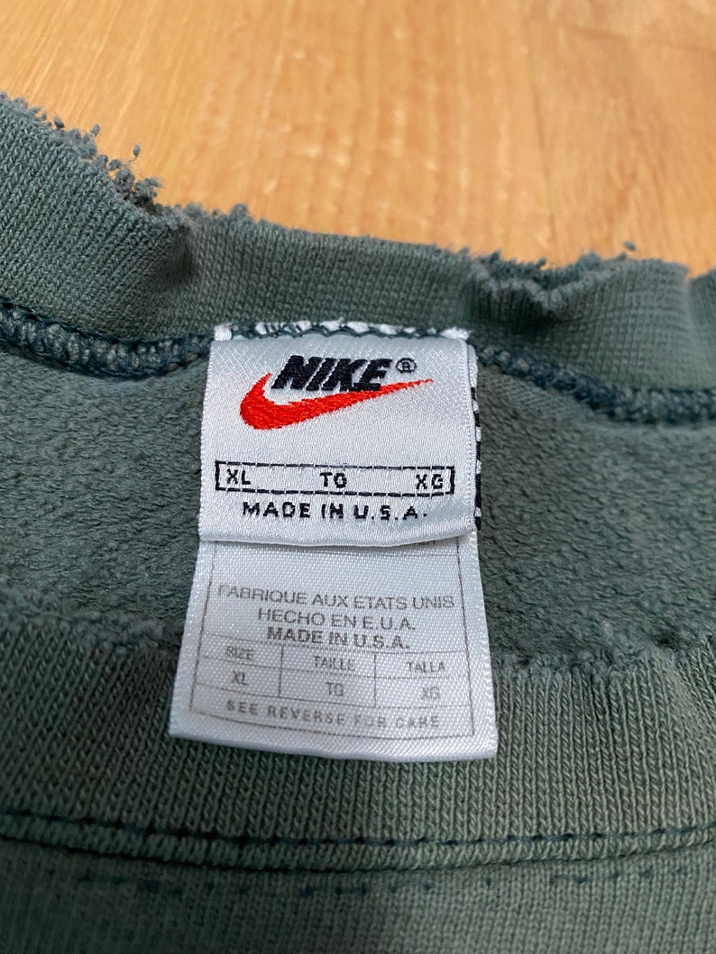 May include: A green Nike sweatshirt tag with the text "XL TO XG MADE IN U.S.A." and "FABRIQUE AUX ETATS UNIS HECHO EN E.U.A. MADE IN U.S.A.". The tag also has a size chart with the sizes "XL", "TG", and "XG".
